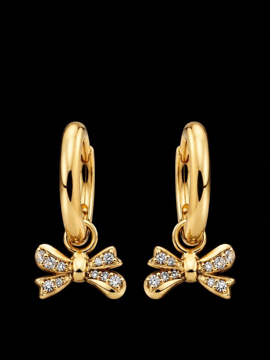 Pair of gold-plated huggie hoop earrings with pavé cubic zirconia bow charms on black background