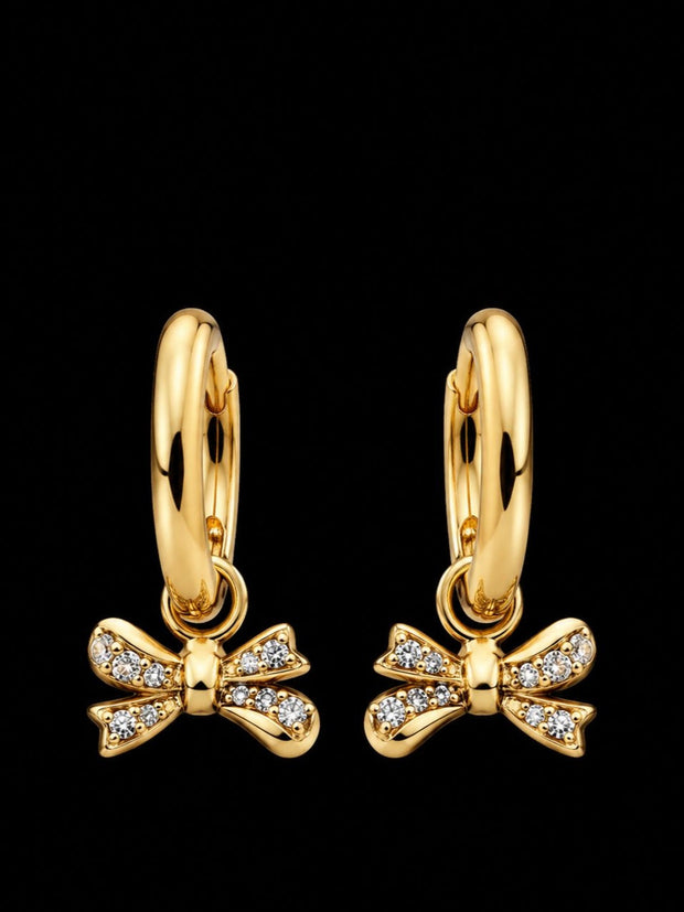 Pair of gold-plated huggie hoop earrings with pavé cubic zirconia bow charms on black background