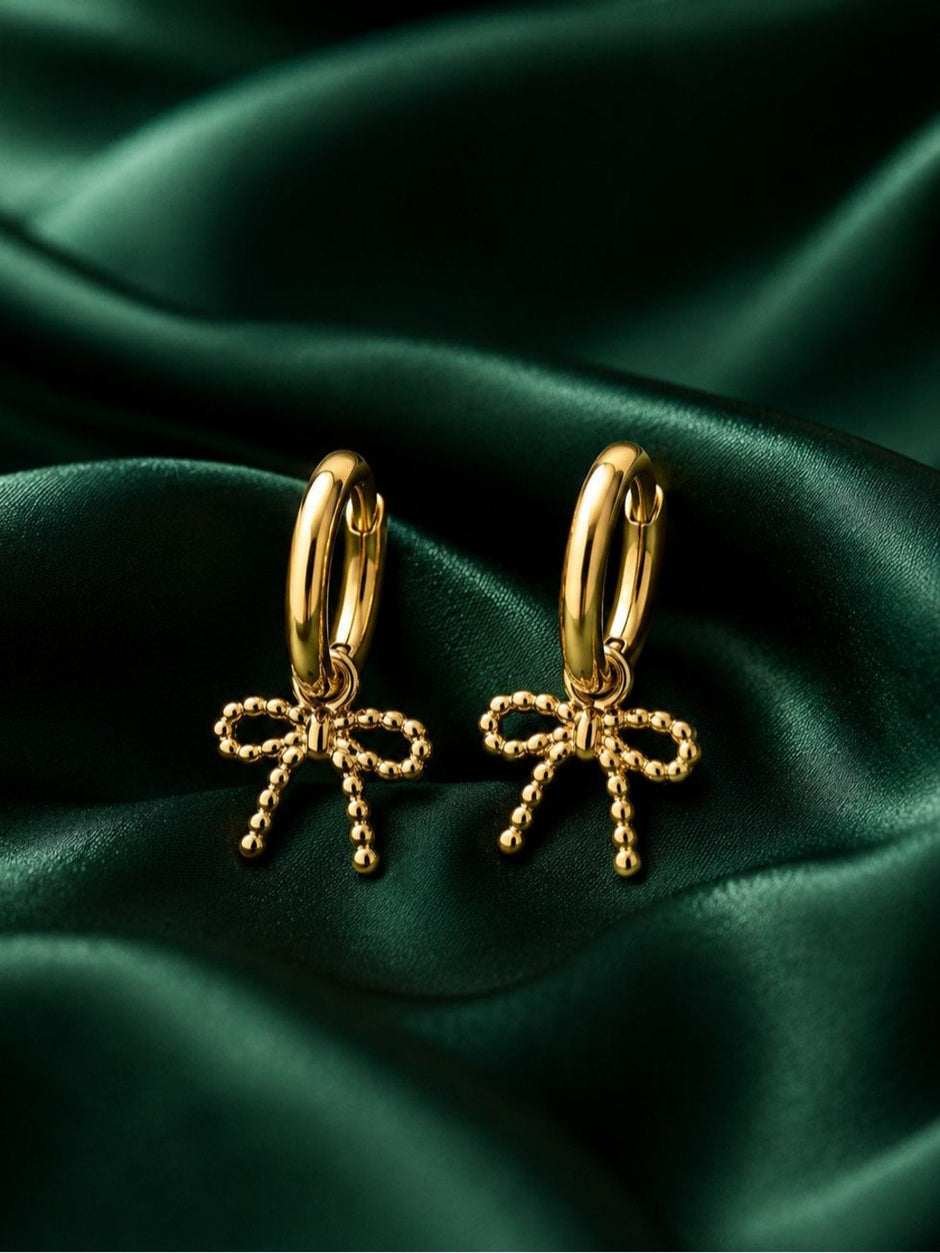 Pair of gold-plated hoop earrings with beaded bow charms laid on emerald green satin fabric