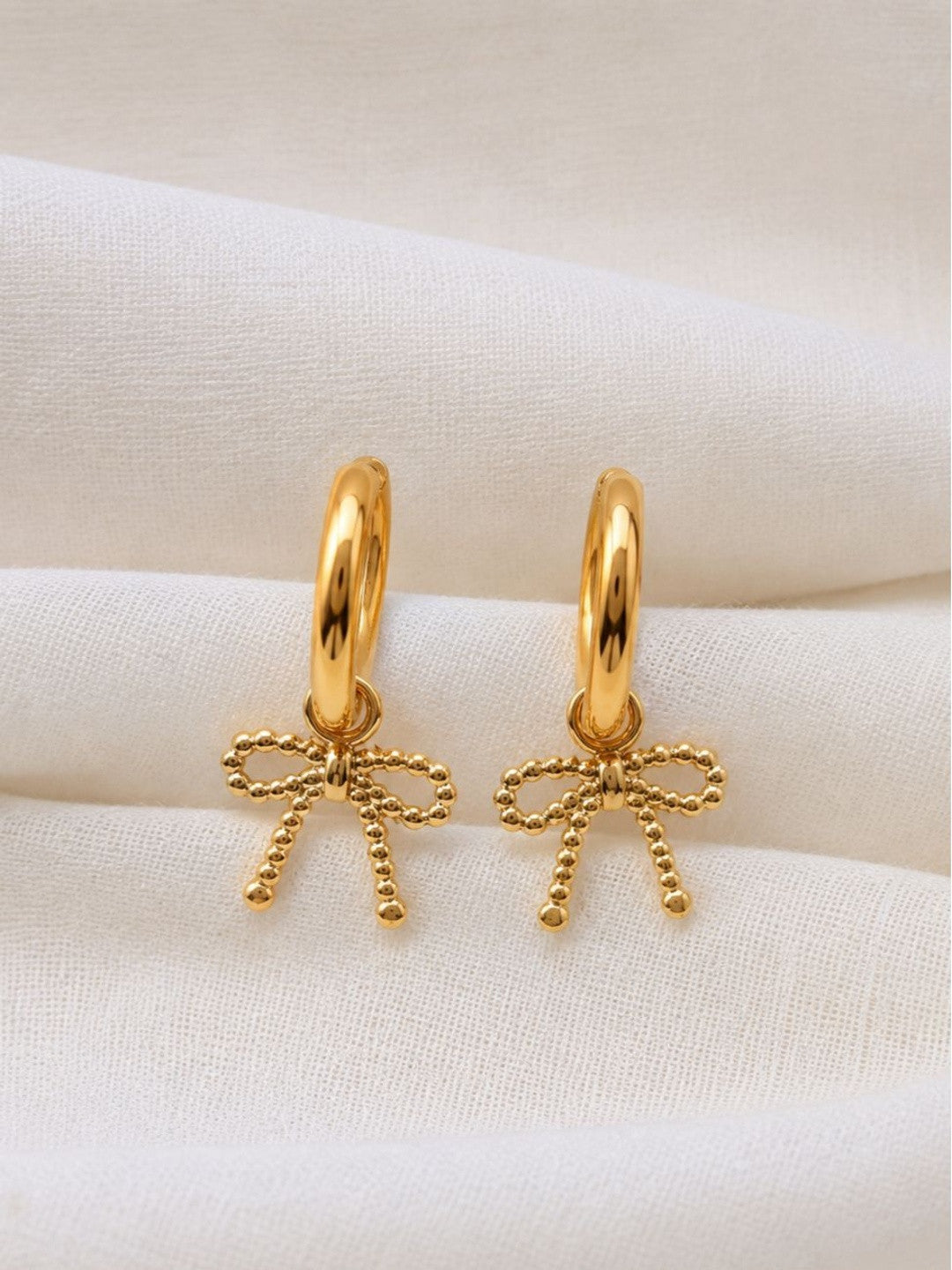 Close-up of two gold bow charm hoop earrings resting on white cloth showing beaded bow detail