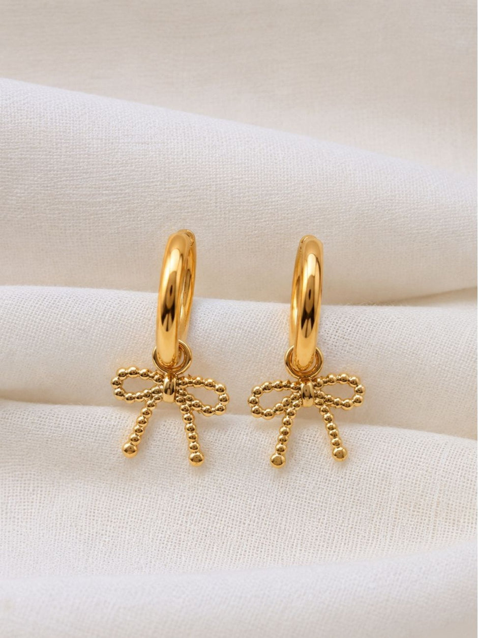 Close-up of two gold bow charm hoop earrings resting on white cloth showing beaded bow detail