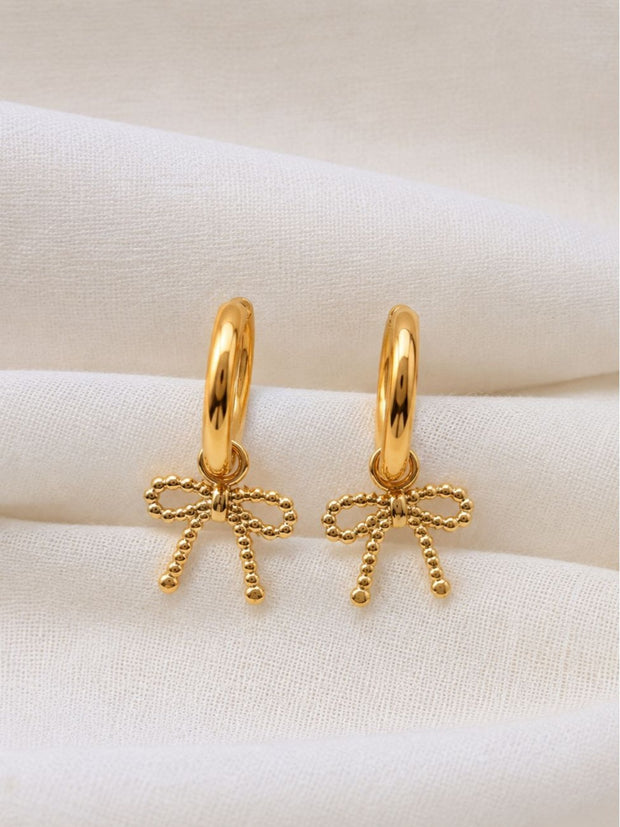 Close-up of two gold bow charm hoop earrings resting on white cloth showing beaded bow detail