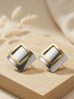 Two-tone geometric square stud earrings placed on a light wooden display block with dried pampas grass in background