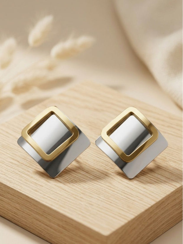 Two-tone geometric square stud earrings placed on a light wooden display block with dried pampas grass in background