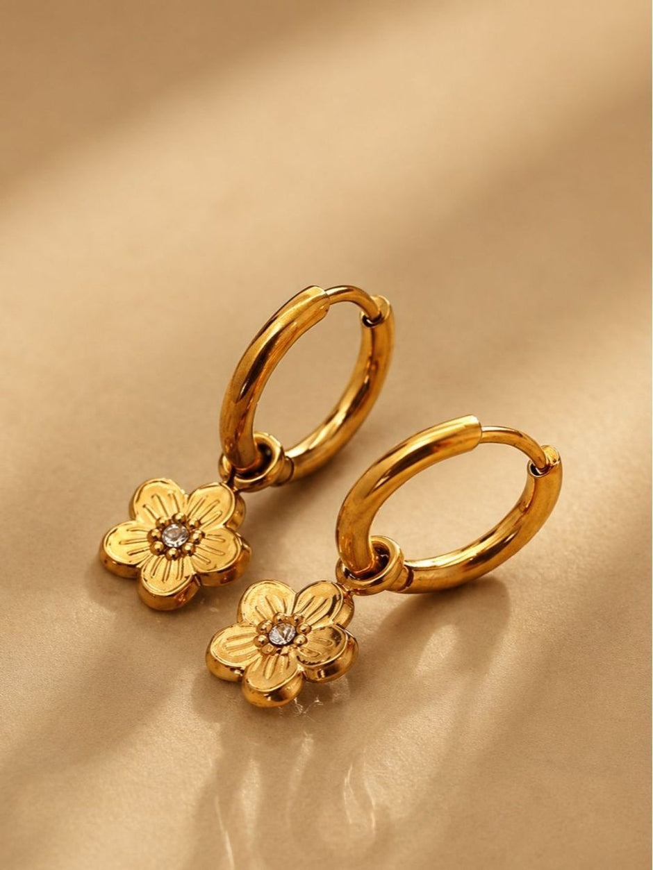 Pair of gold-plated flower charm hoop earrings with engraved petals and crystal centre on beige background