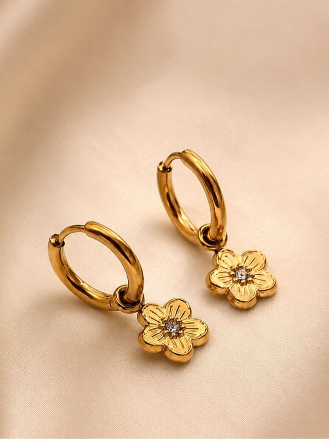 Two gold-plated floral hoop earrings laid flat on a cream satin fabric surface