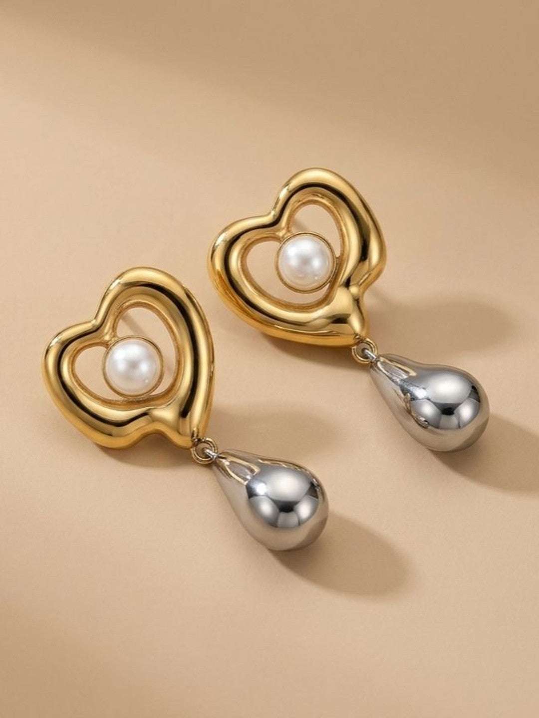 Pair of gold-tone open-heart earrings with faux pearl centre and silver teardrop pendants on beige surface