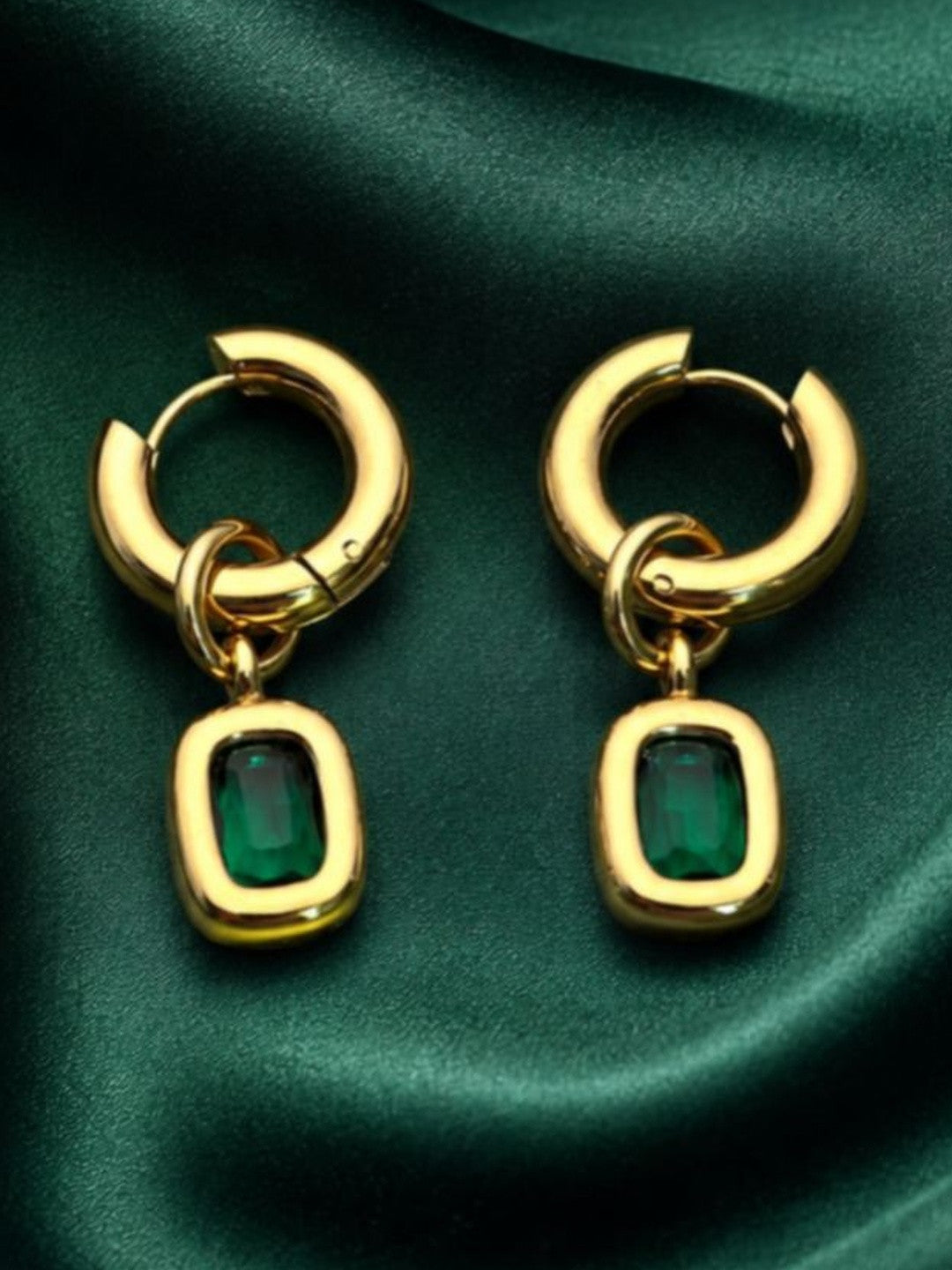Pair of gold-plated emerald green cushion drop hoop earrings laid flat on dark green satin fabric