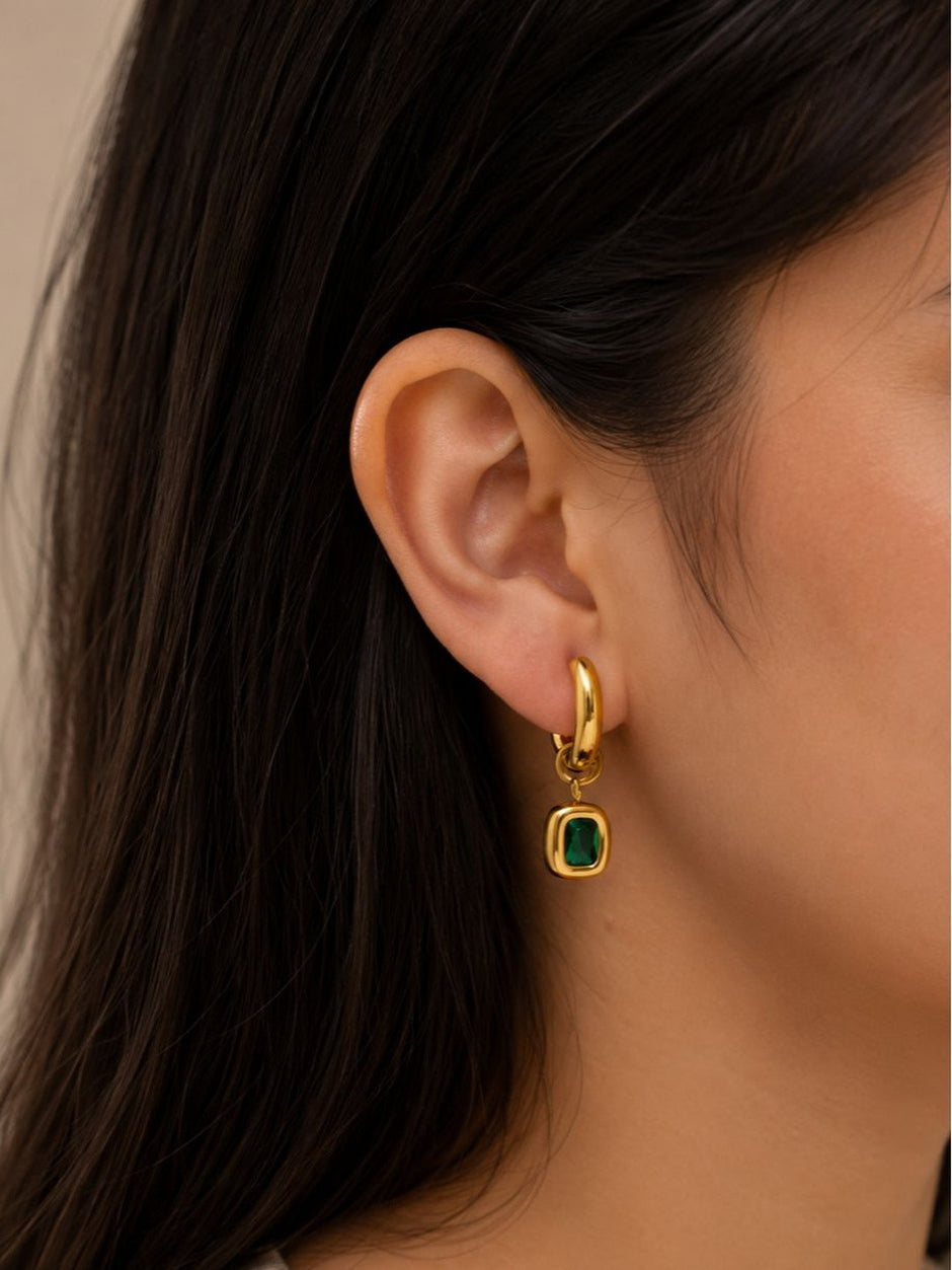Gold-plated emerald green drop hoop earring worn on a womans ear with dark hair in the background