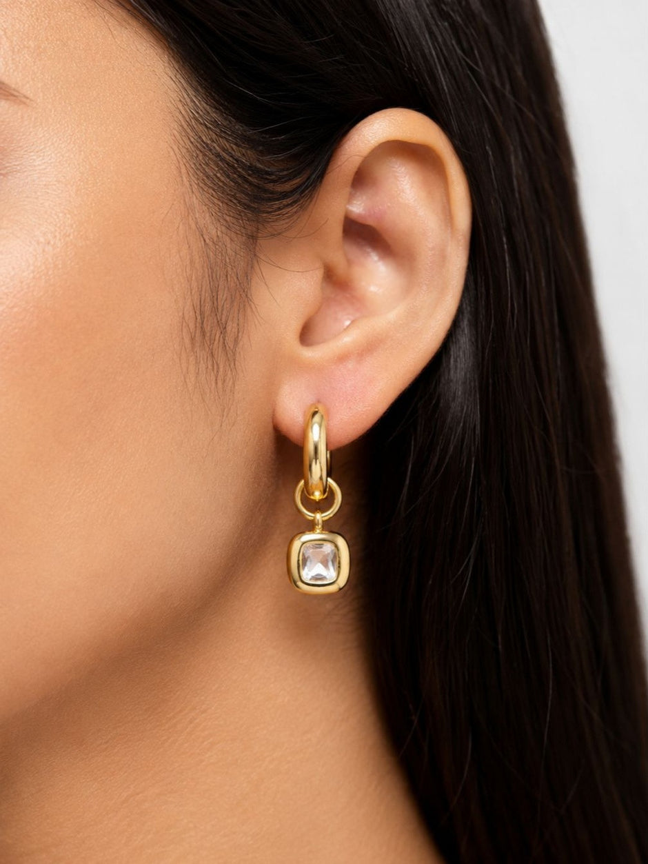 Close-up of gold hoop earring with cushion-cut crystal charm pendant worn on a woman's ear