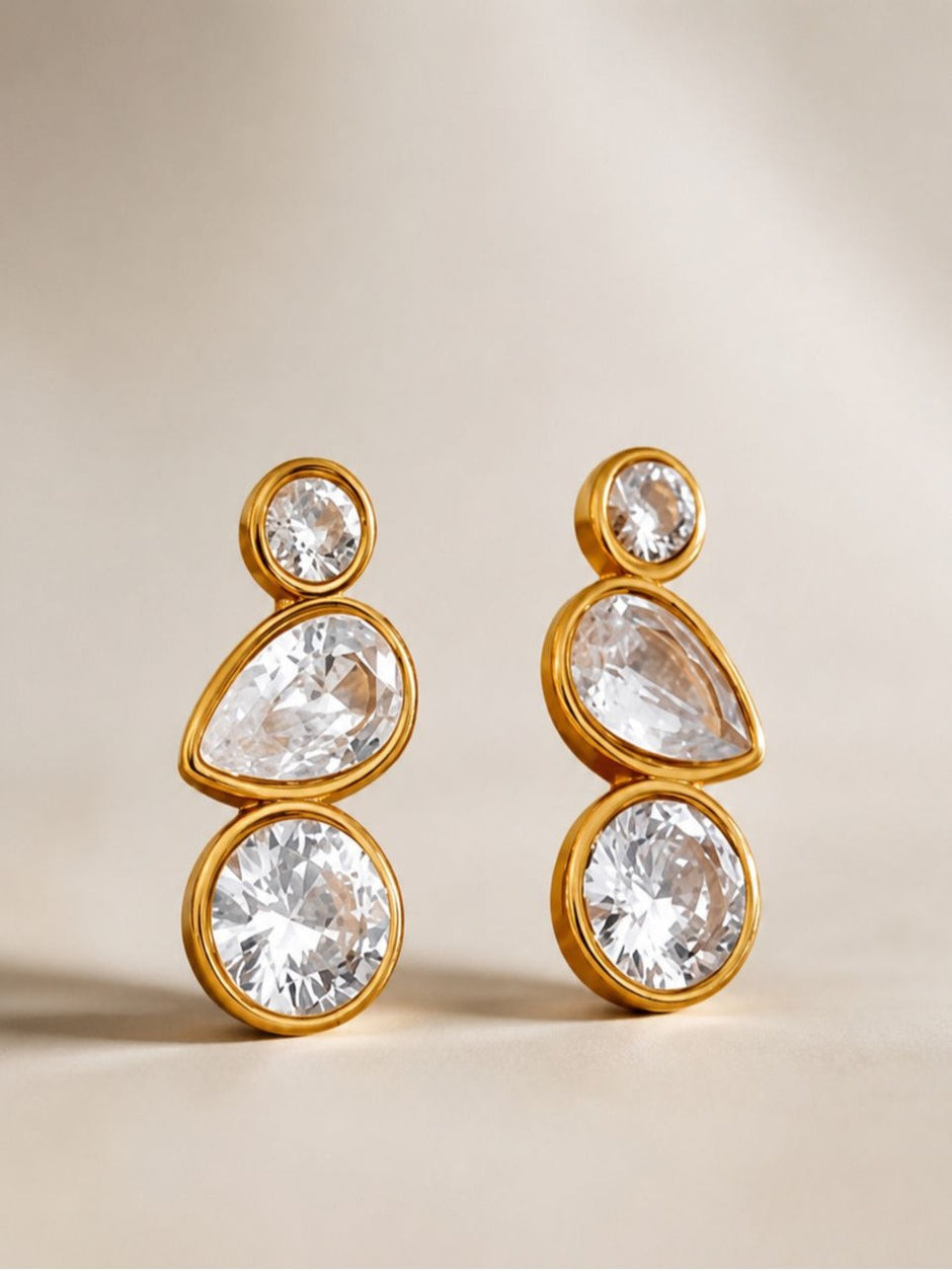 Front view of gold-plated crystal bezel drop earrings standing upright on a beige surface
