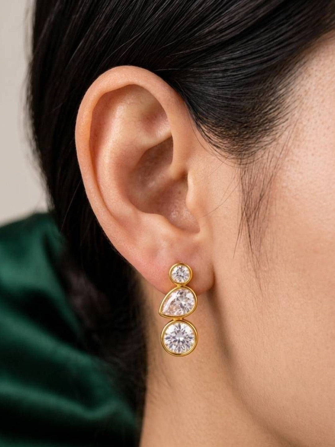 Gold-plated bezel drop earring with three crystal stones worn by a dark-haired model against green fabric