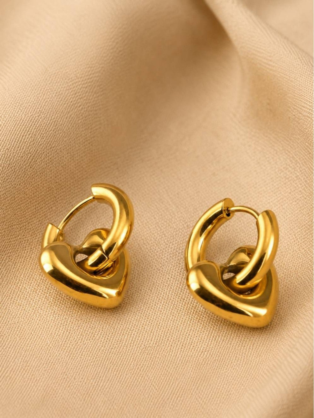 Gold heart charm hoop earrings lying flat on beige fabric showing hinge closure detail