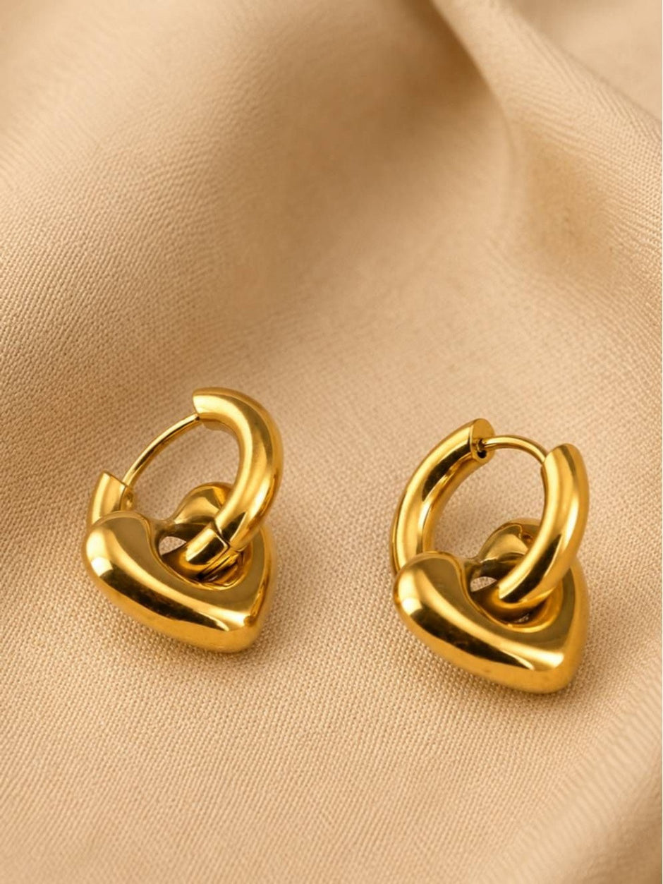 Gold heart charm hoop earrings lying flat on beige fabric showing hinge closure detail