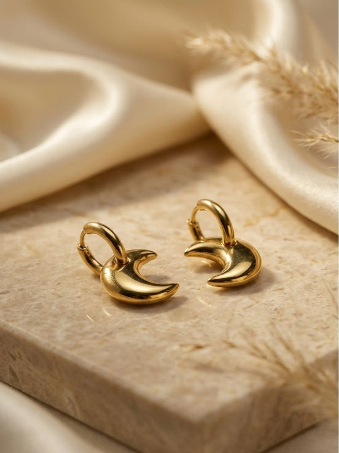Pair of gold-plated crescent moon hoop earrings resting on a beige stone surface with cream fabric