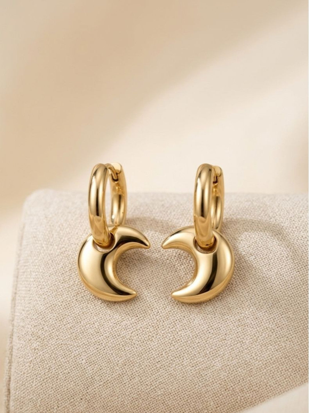 Two gold crescent moon charm hoops placed facing each other on a linen surface