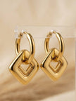 Gold-plated hoop earrings with diagonal square drop pendants displayed upright on a clear acrylic earring stand