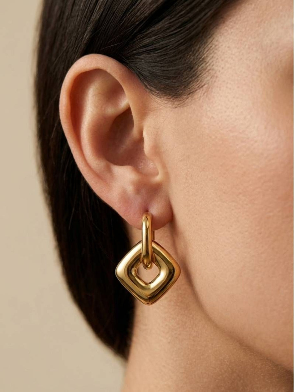 Close-up side view of gold geometric square drop earring worn on a model with dark straight hair