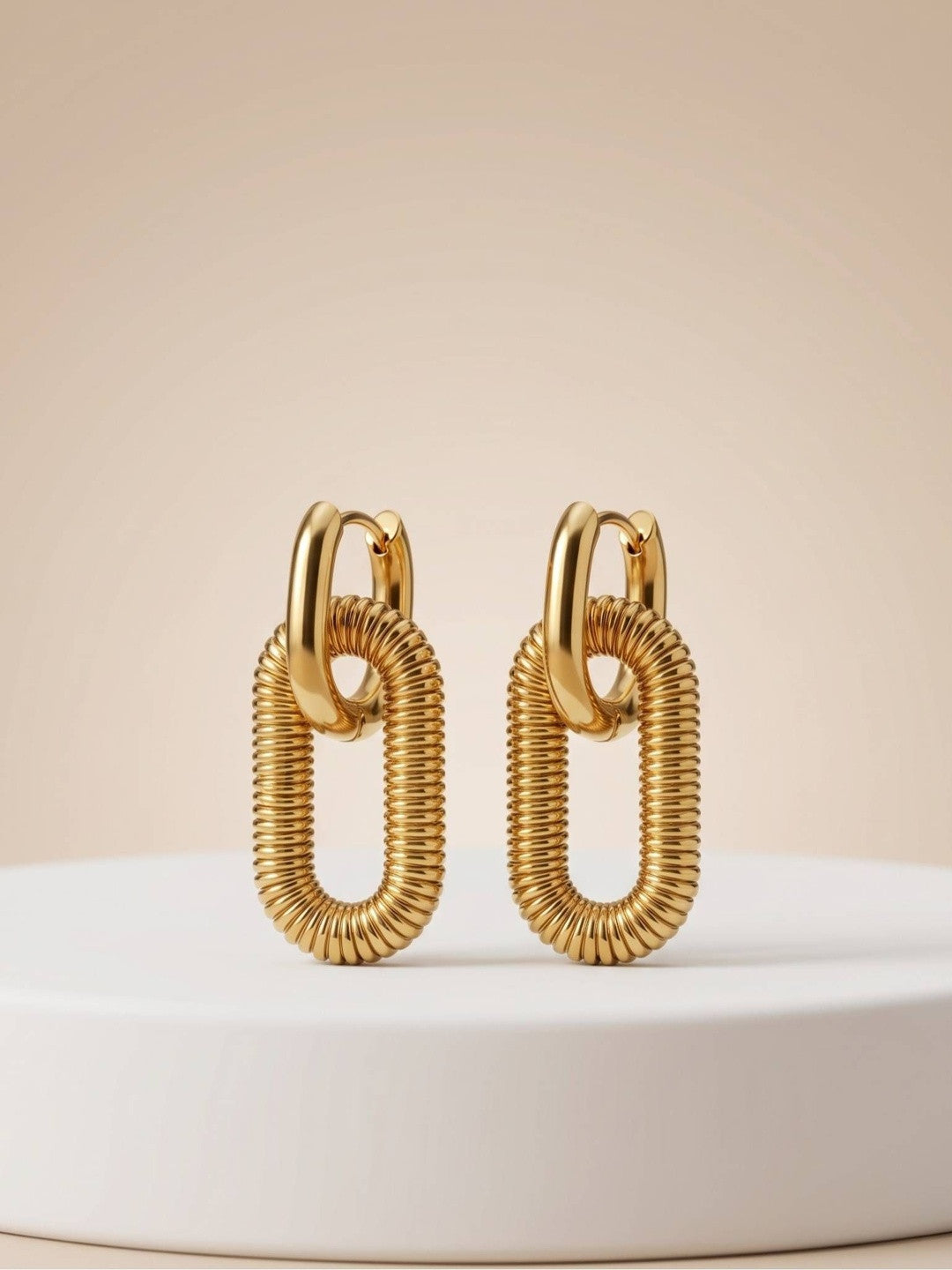 Pair of gold-plated ribbed oval link drop earrings standing upright on a white display pedestal