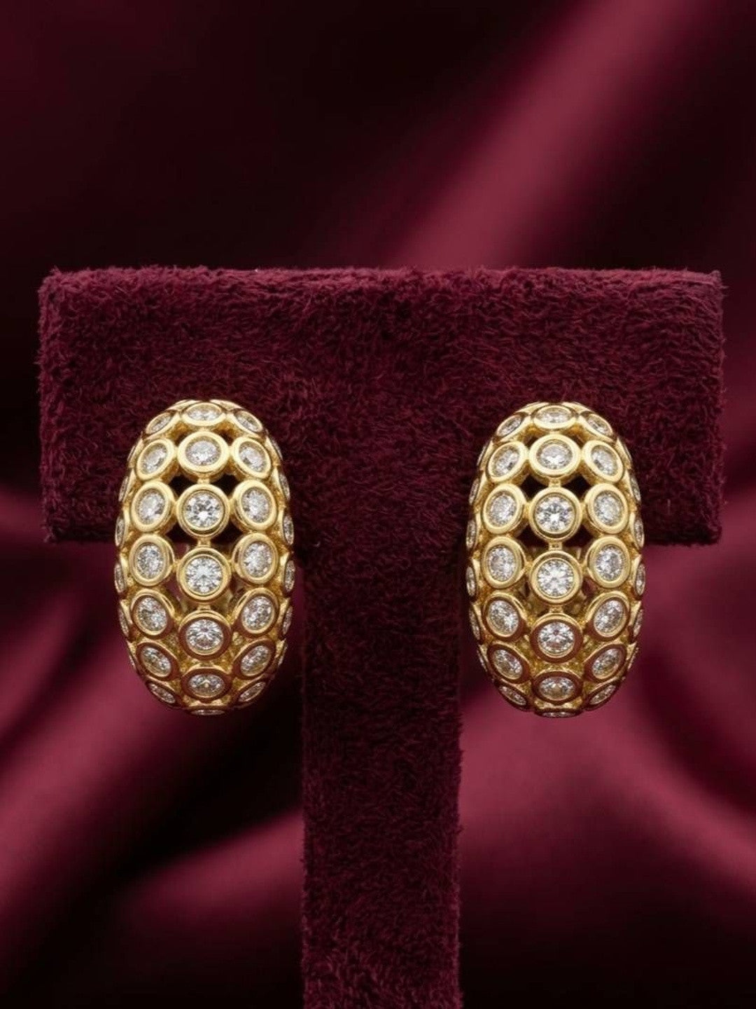 Pair of gold-plated dome hoop earrings covered in bezel-set crystal stones displayed on burgundy velvet earring stand
