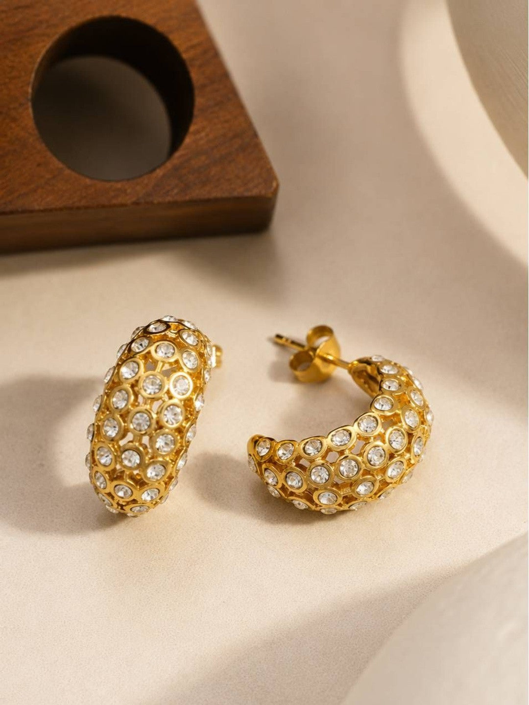 Close-up of two gold crystal bezel hoop earrings with post backs resting on cream surface near wooden prop