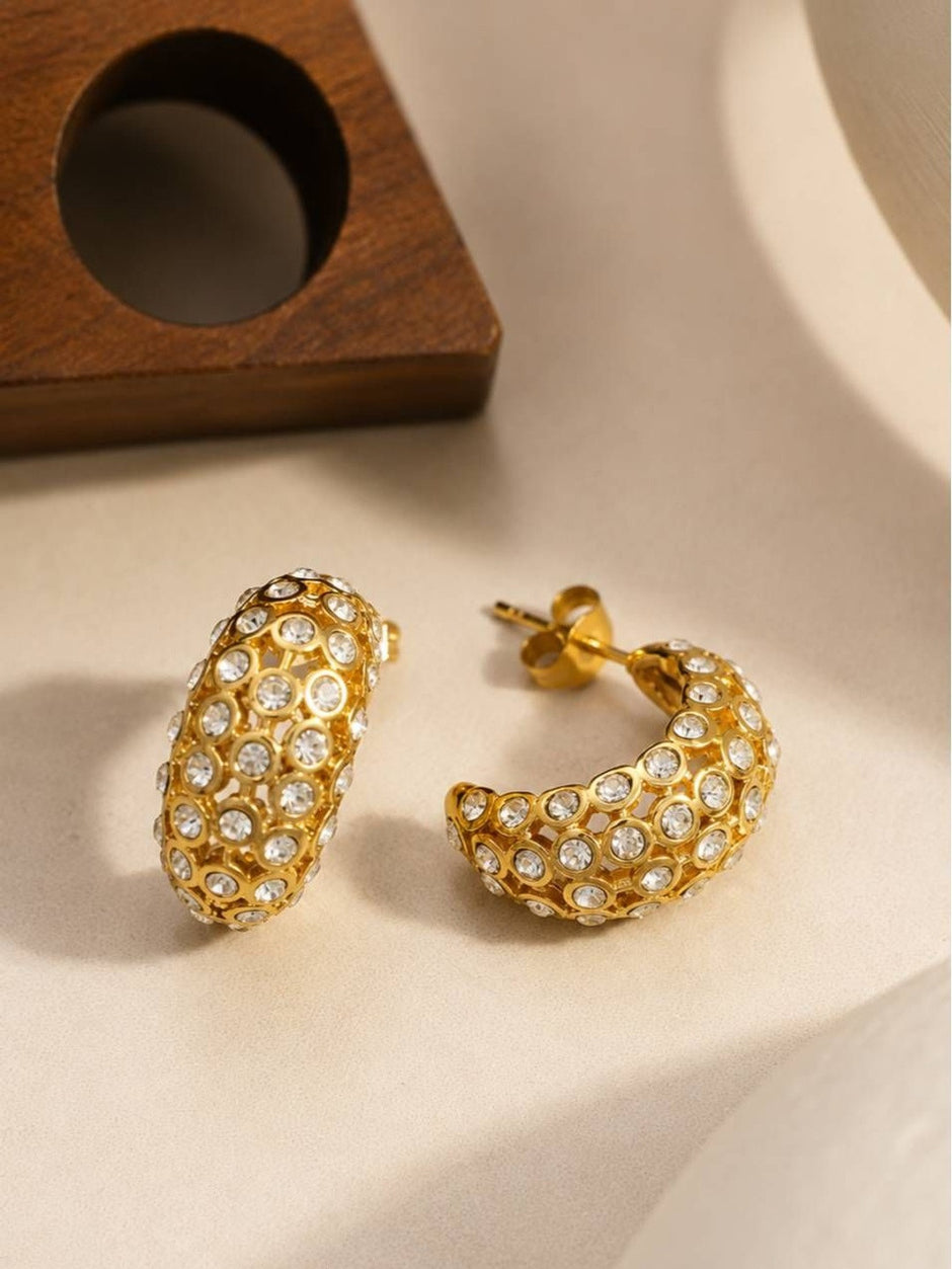 Close-up of two gold crystal bezel hoop earrings with post backs resting on cream surface near wooden prop
