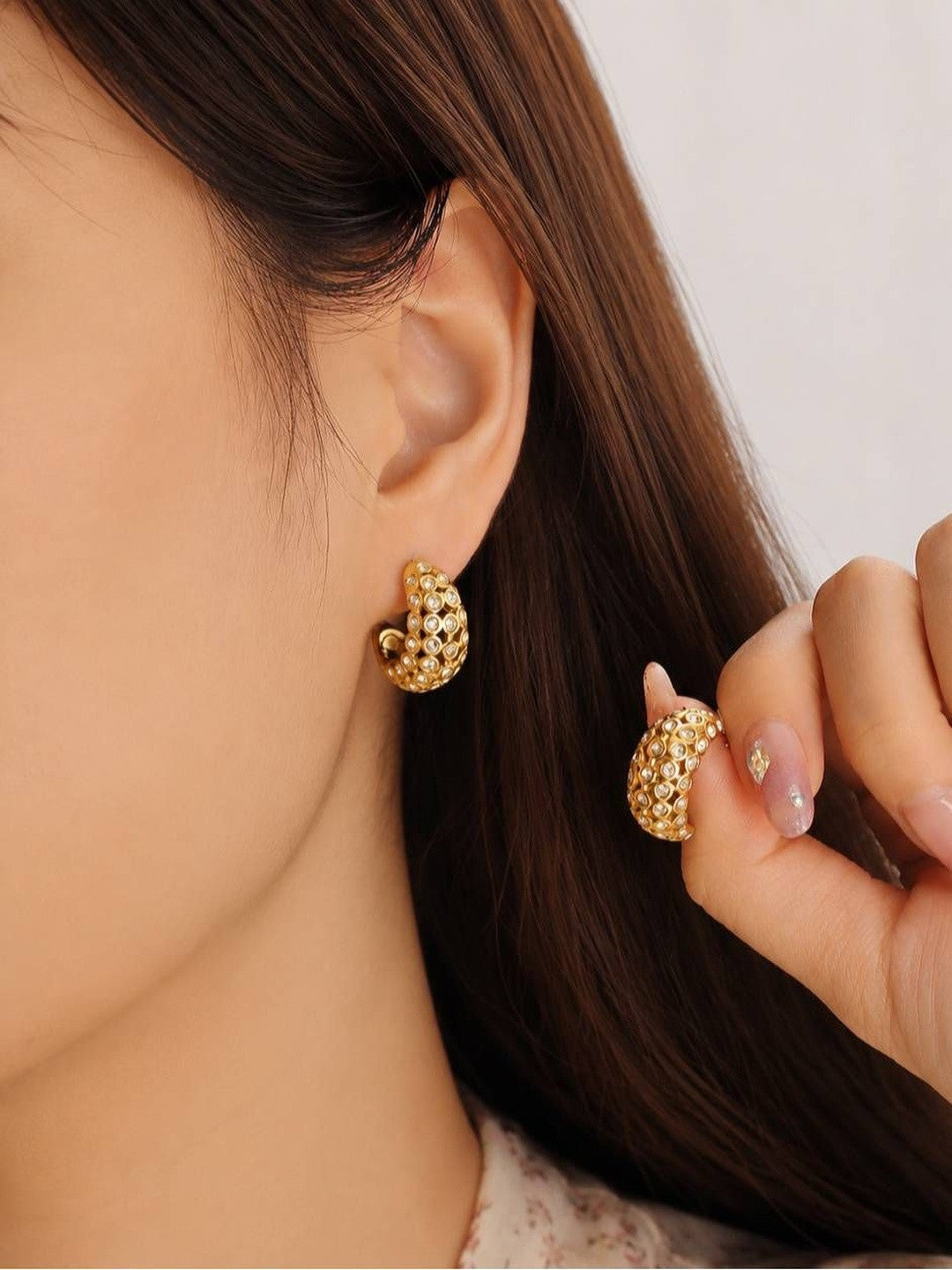 Woman wearing gold crystal dome hoop earring on left ear holding second earring in right hand