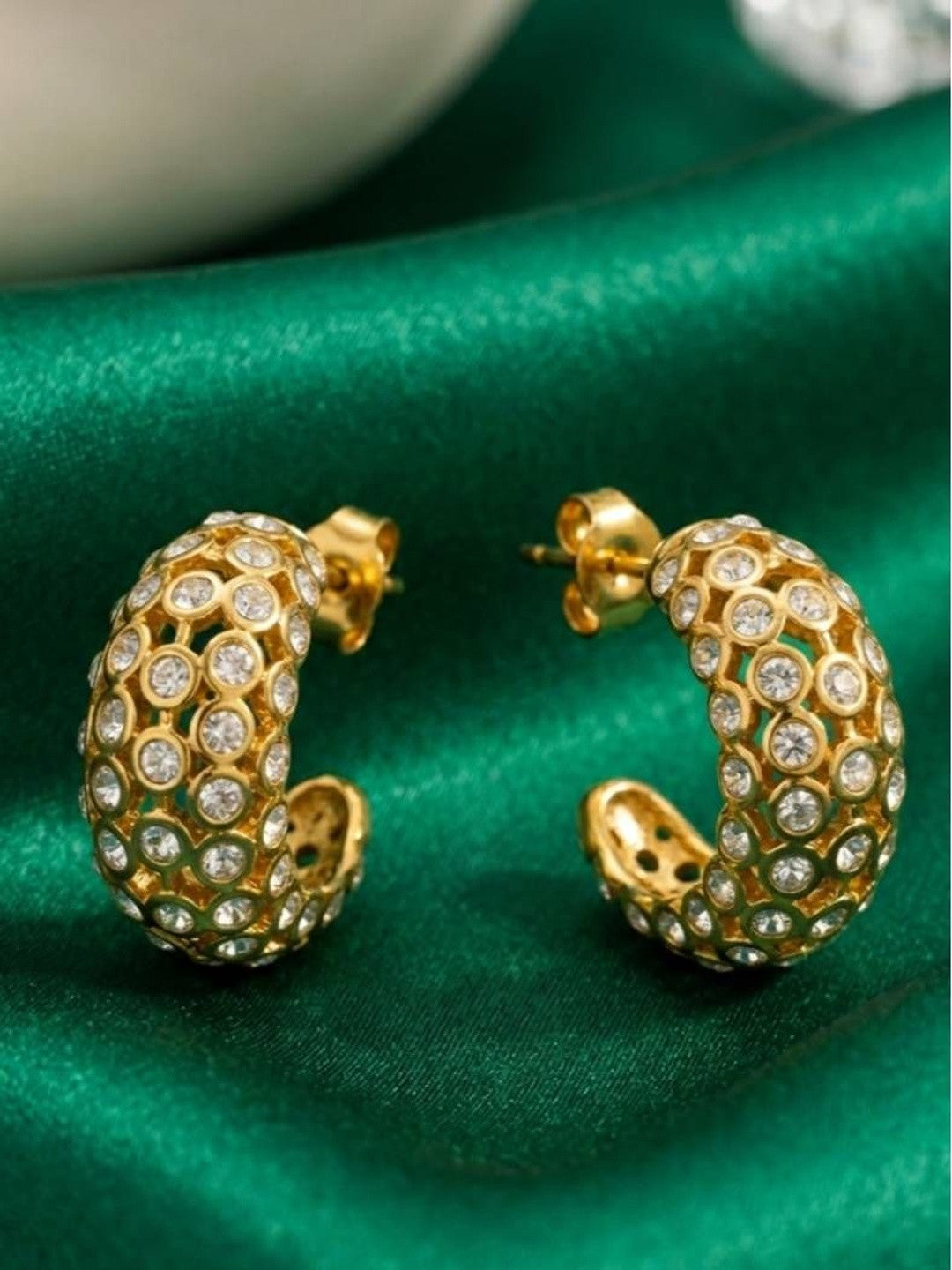 Two gold-plated crystal bezel hoop earrings placed on emerald green satin fabric showing open lattice underside