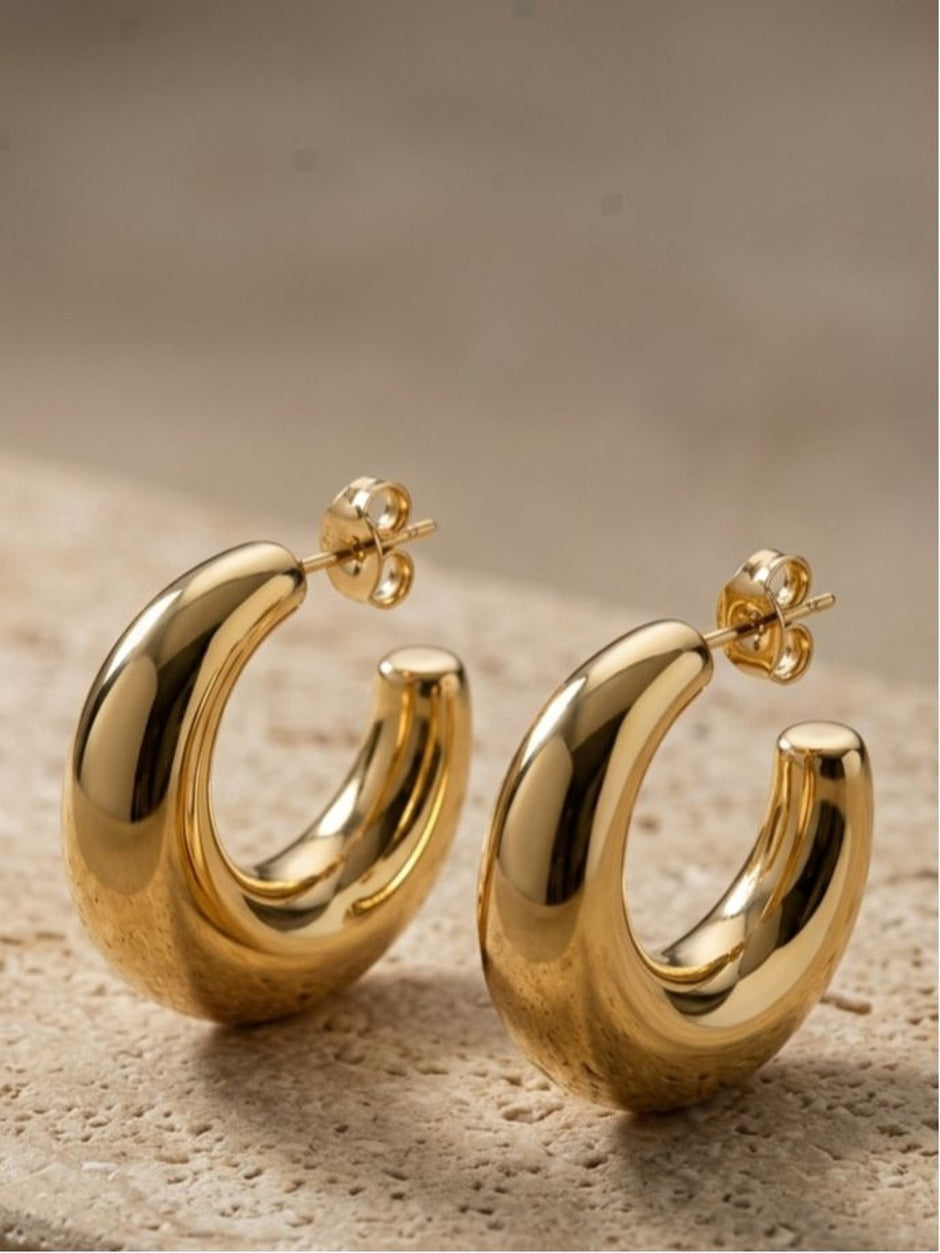 Pair of chunky gold-plated oval half hoop earrings with push-back posts resting on a textured stone surface