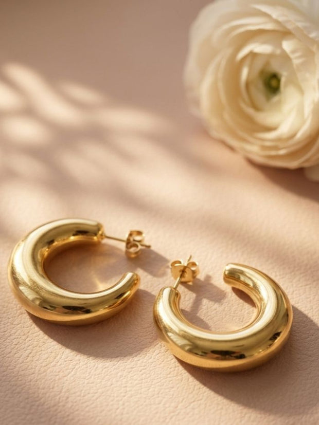 Two gold-plated chunky oval half hoop earrings lying flat on a blush surface beside a white ranunculus flower