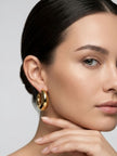 Front-facing model with dark hair pulled back wearing chunky gold-plated oval half hoop earrings, hand resting near chin