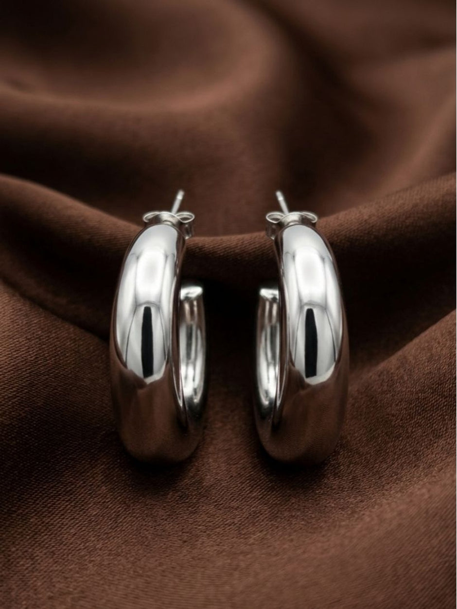 Pair of chunky silver-finish hoop earrings laid flat on rich brown fabric