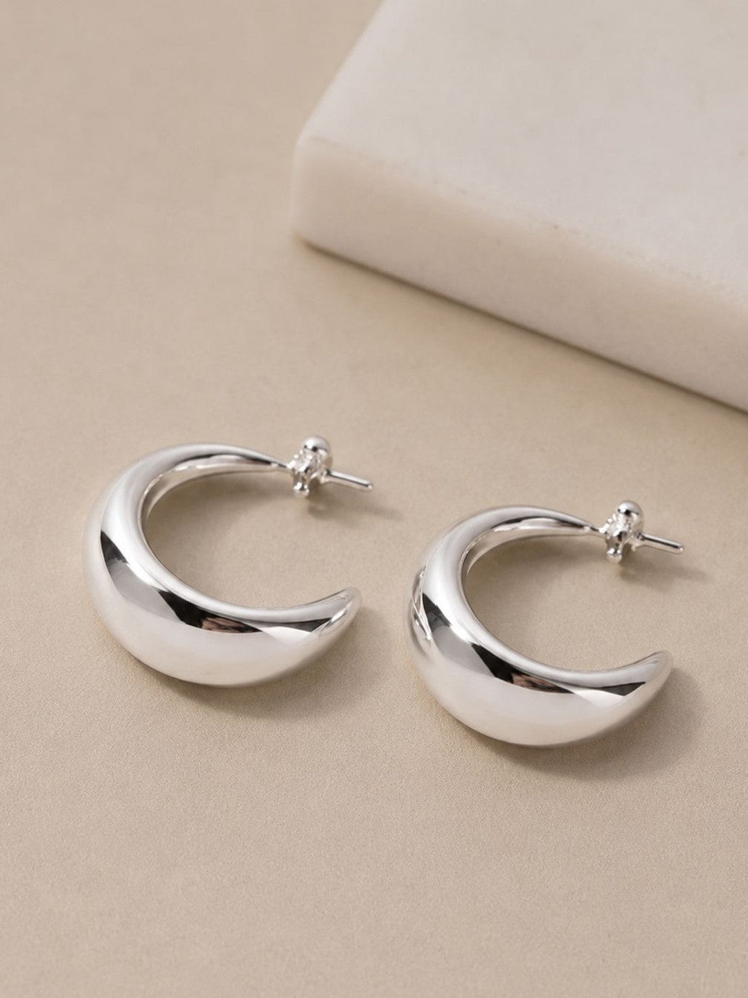 Two chunky silver C-hoop earrings with crystal-set posts resting on a cream surface