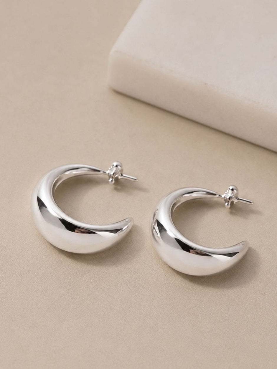Two chunky silver C-hoop earrings with crystal-set posts resting on a cream surface