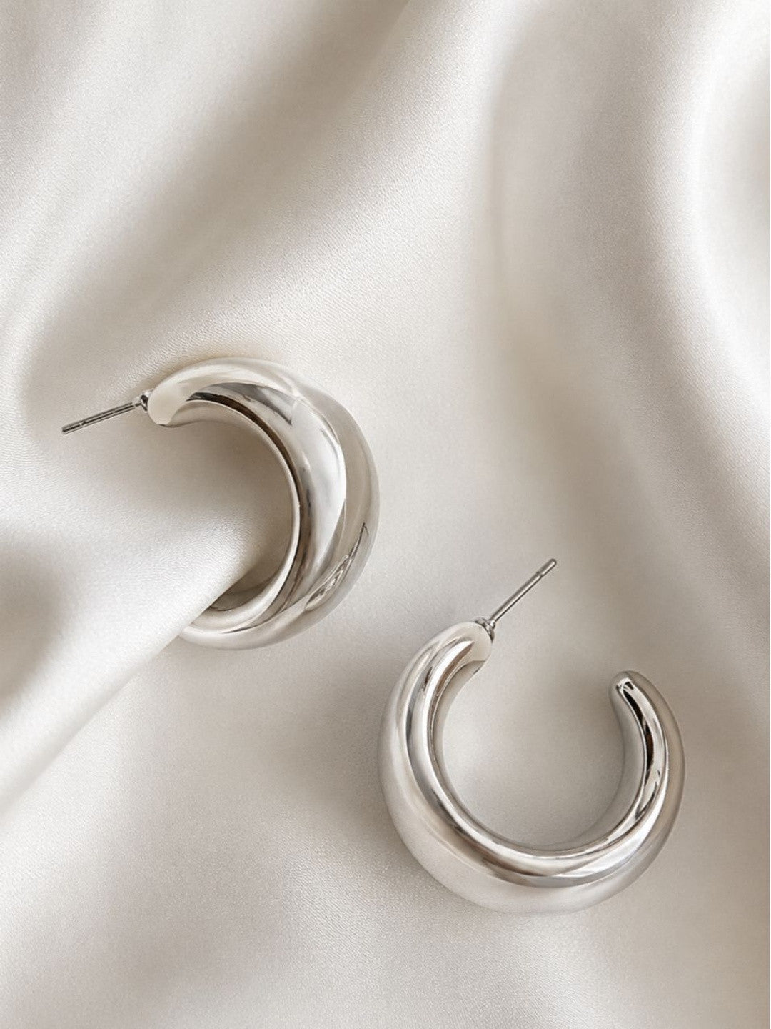 Pair of chunky silver tubular hoop earrings placed on draped white satin fabric