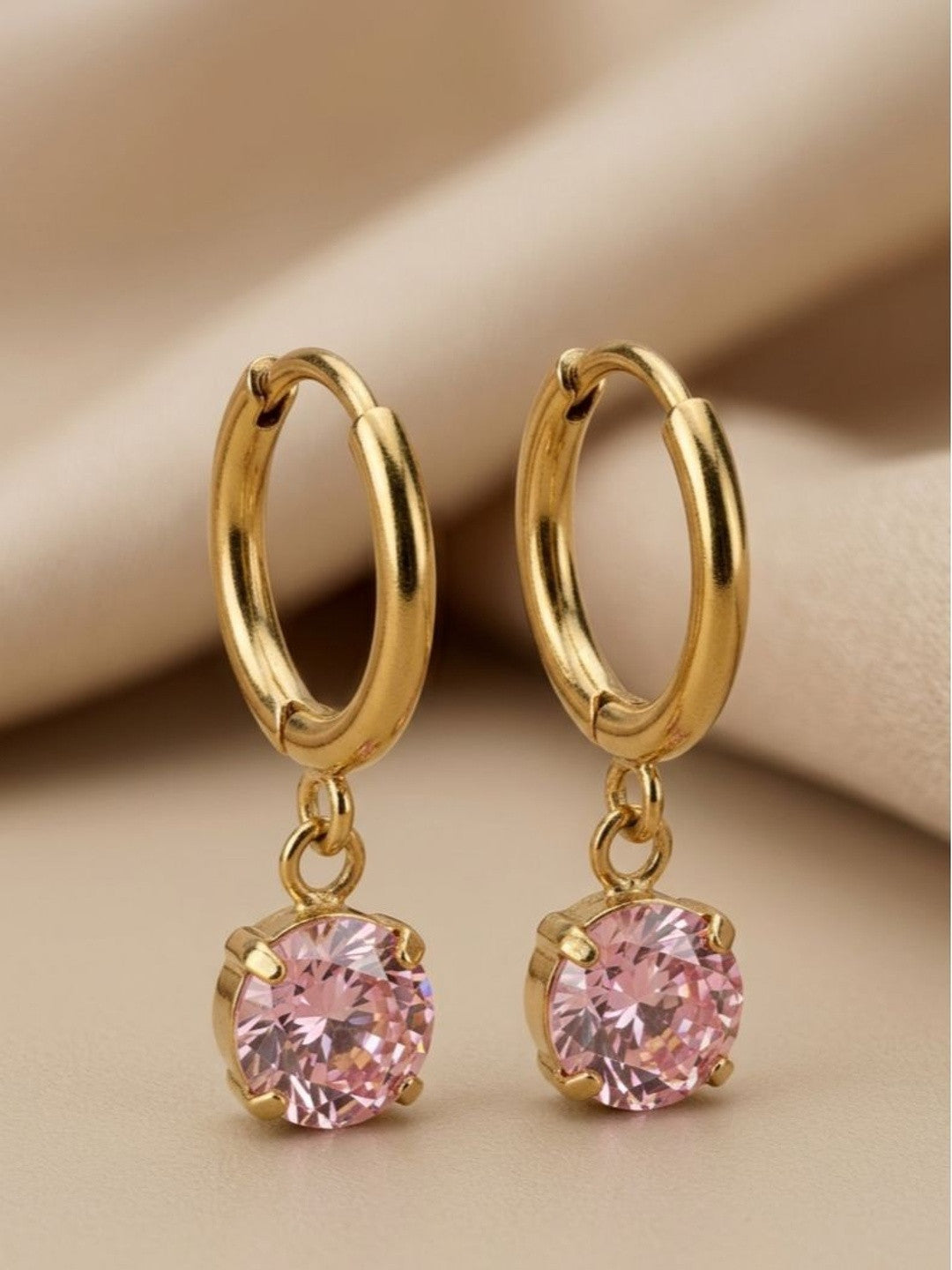 Close-up of gold huggie hoops with pink solitaire charm dangle on beige fabric