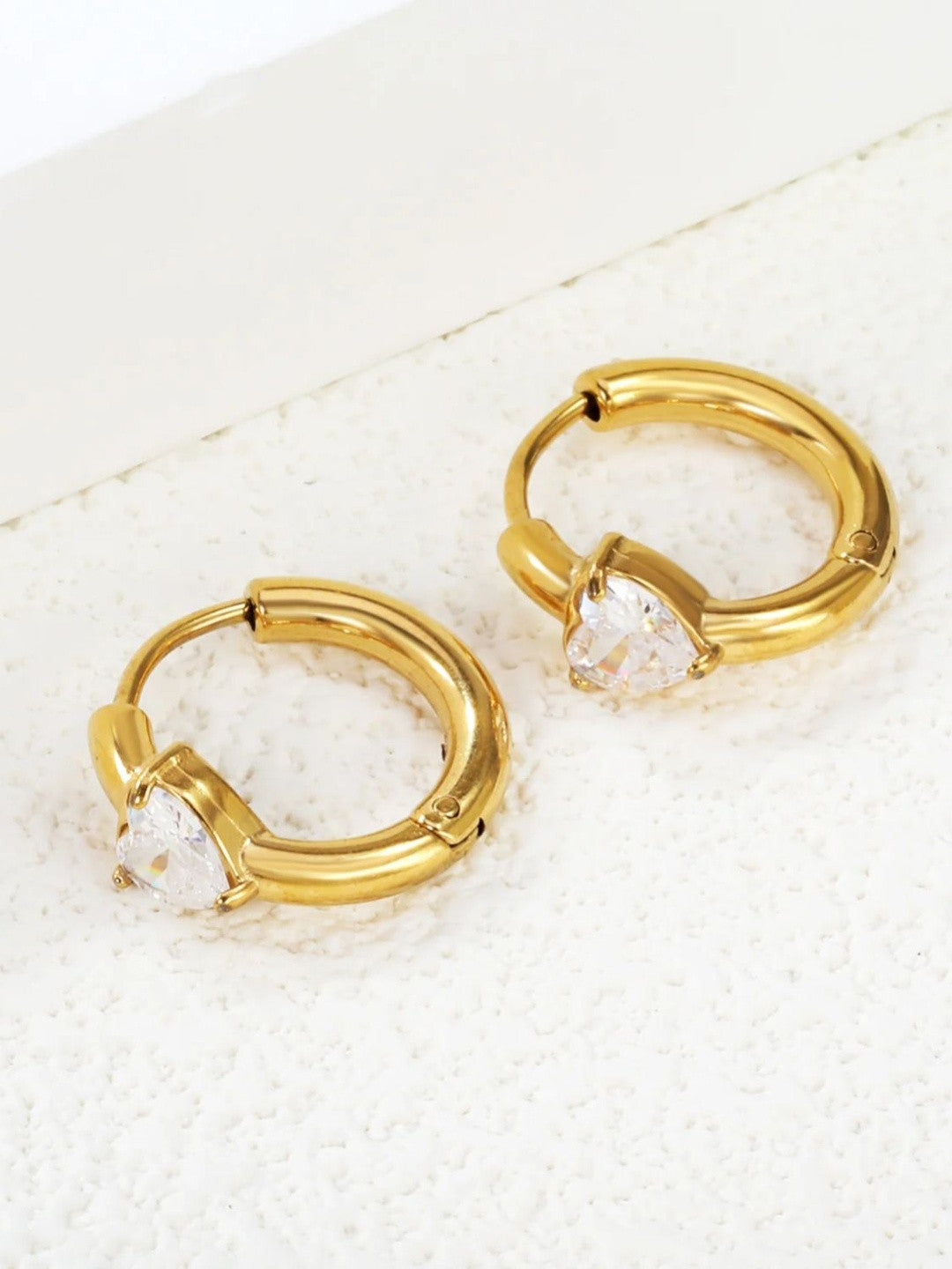 Two gold-plated heart CZ huggie hoop earrings laid flat on white textured surface