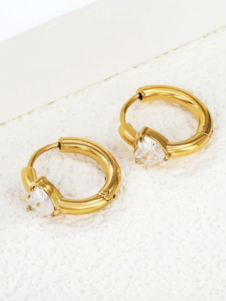 Two gold-plated heart CZ huggie hoop earrings laid flat on white textured surface