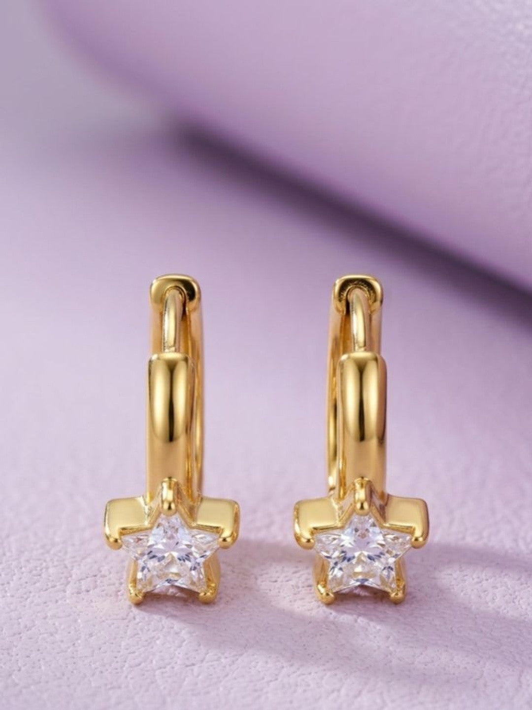 Close-up of two gold-plated star cubic zirconia hoop earrings against a soft lavender background