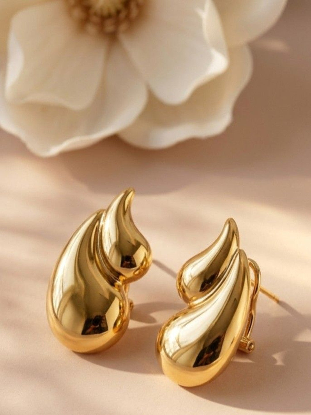 Pair of gold-toned chunky flame drop stud earrings resting on a beige surface beside a white magnolia flower