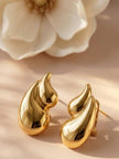 Pair of gold-toned chunky flame drop stud earrings resting on a beige surface beside a white magnolia flower