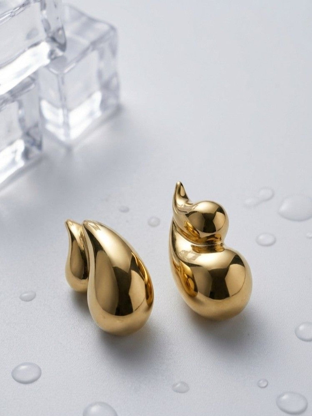 Two gold-toned flame drop stud earrings shown from different angles on a wet grey surface beside ice cubes