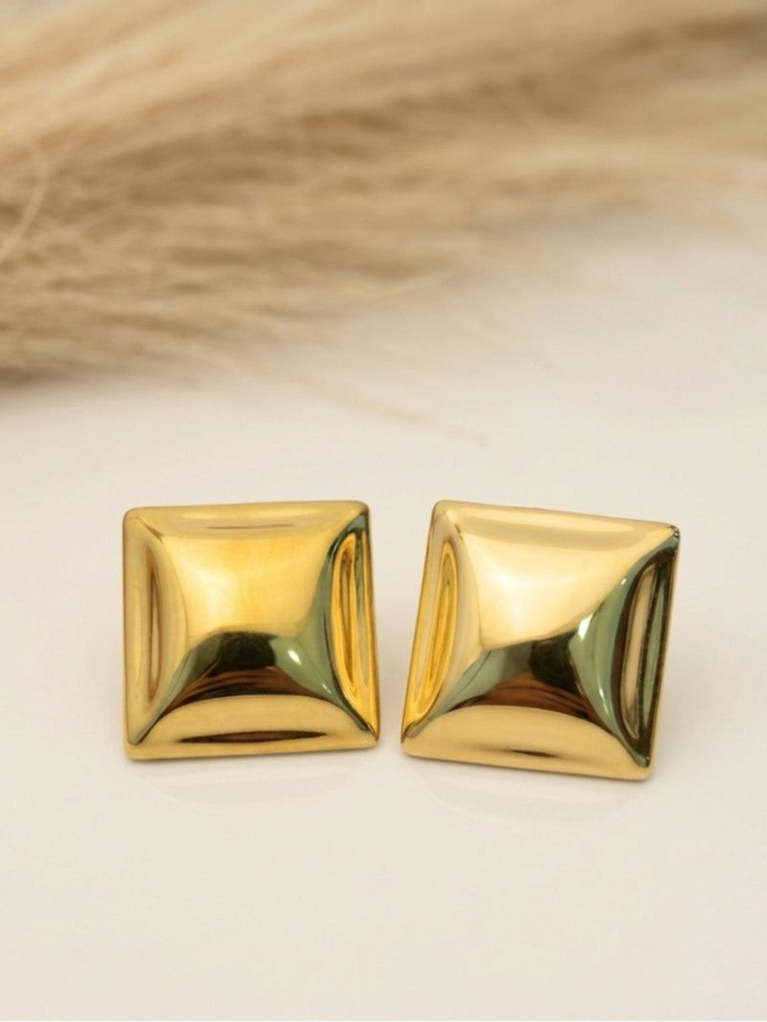 Pair of gold-plated square pyramid studs on white surface with dried grass backdrop