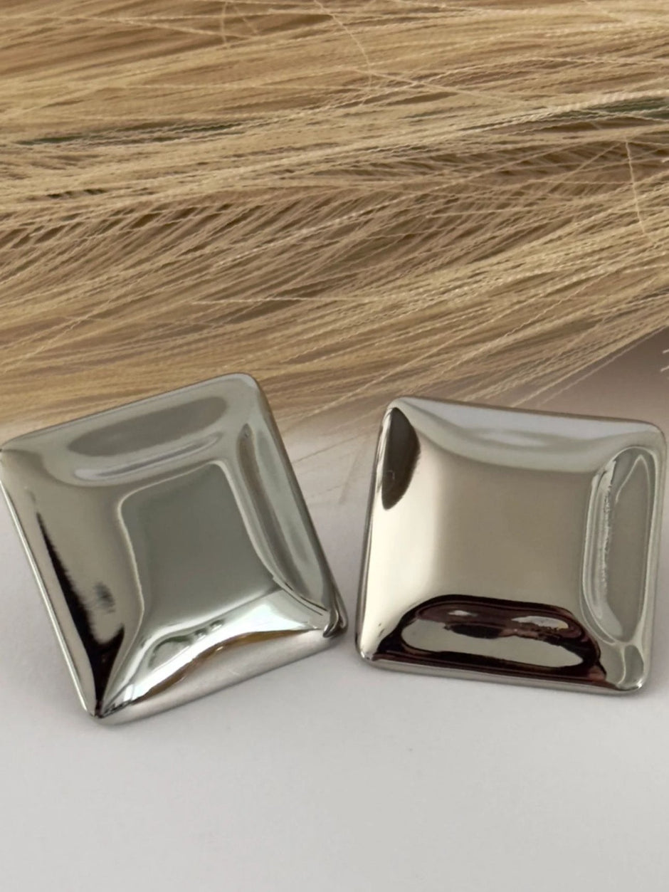 Pair of silver-finish cushion square stud earrings laid flat on a white surface with blonde hair in background