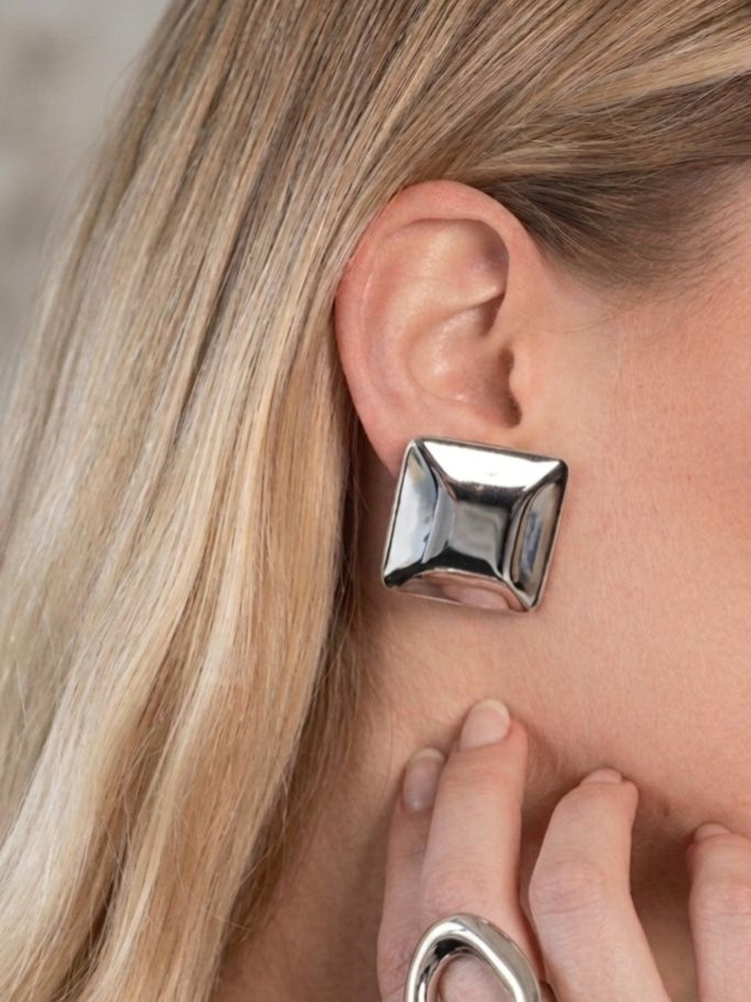 Silver square stud earring on the ear of a blonde model, hand resting near jaw with matching silver ring visible