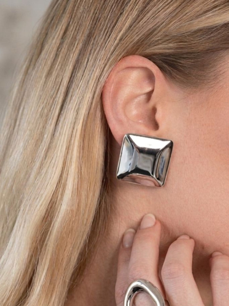 Silver square stud earring on the ear of a blonde model, hand resting near jaw with matching silver ring visible