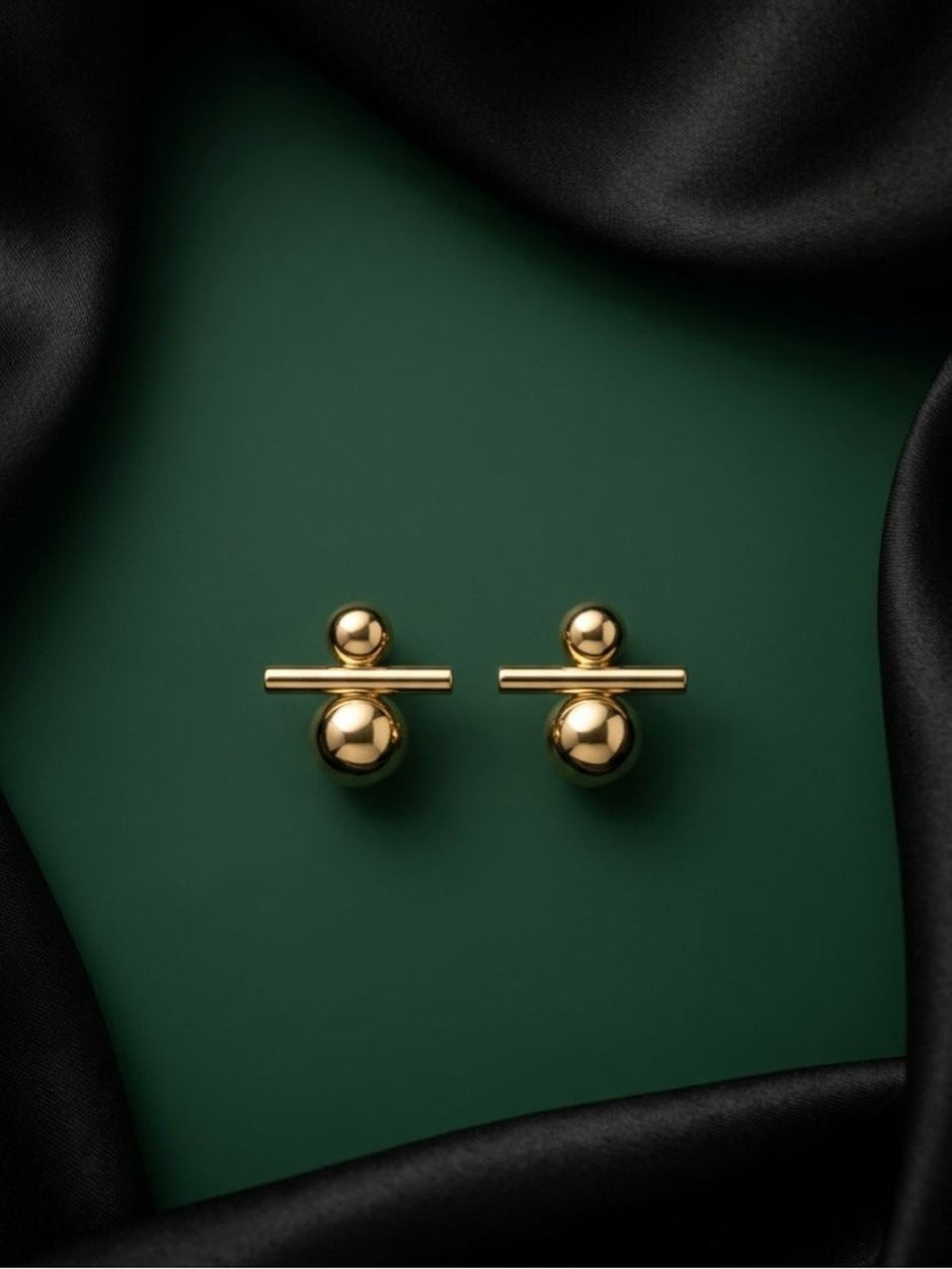 Pair of gold-toned ball and bar drop earrings laid flat on a dark green surface with black satin fabric