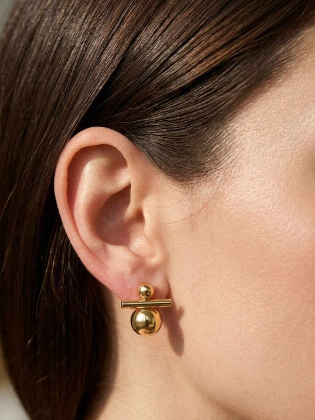 Gold ball and bar drop earring worn on the ear of a woman with dark hair pulled back