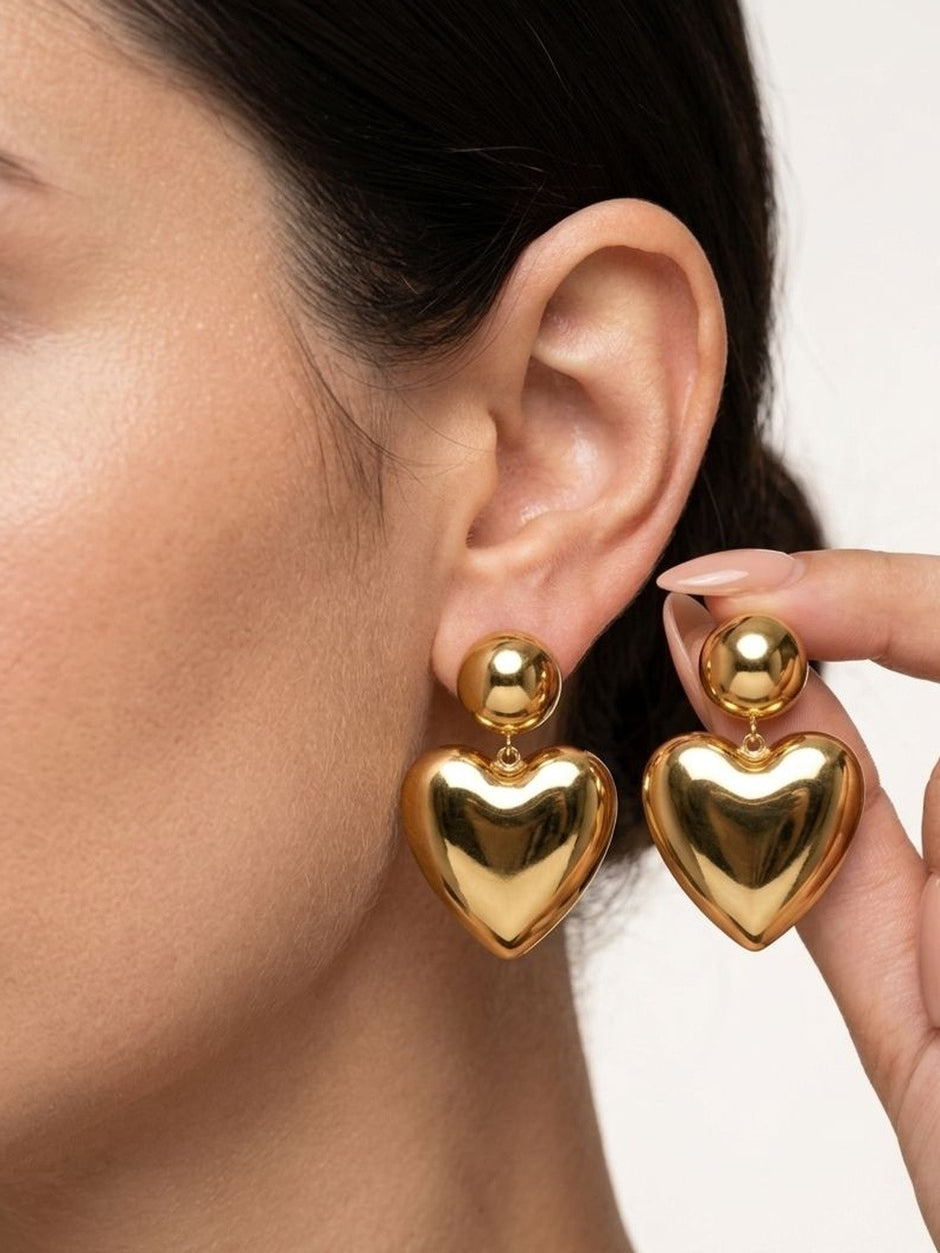 Close-up of gold puffy heart drop earrings worn on a model's ear, hand holding second earring
