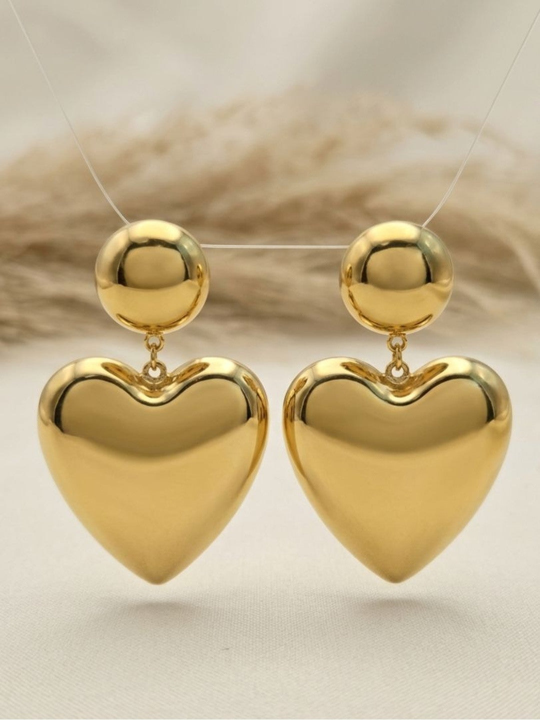Gold heart drop earrings suspended on a display stand against a neutral linen background